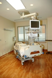 The image depicts a clean and modern hospital room featuring a hospital bed with advanced monitoring equipment attached. The room is well-lit with ceiling lights and has a large window with blinds, allowing natural light to enter. The floors are made of polished wood, and there are neutral-colored walls. Medical equipment and a computer monitor are suspended above the bed, contributing to the sterile and professional atmosphere of the room.