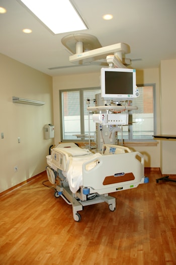 The image depicts a clean and modern hospital room featuring a hospital bed with advanced monitoring equipment attached. The room is well-lit with ceiling lights and has a large window with blinds, allowing natural light to enter. The floors are made of polished wood, and there are neutral-colored walls. Medical equipment and a computer monitor are suspended above the bed, contributing to the sterile and professional atmosphere of the room.