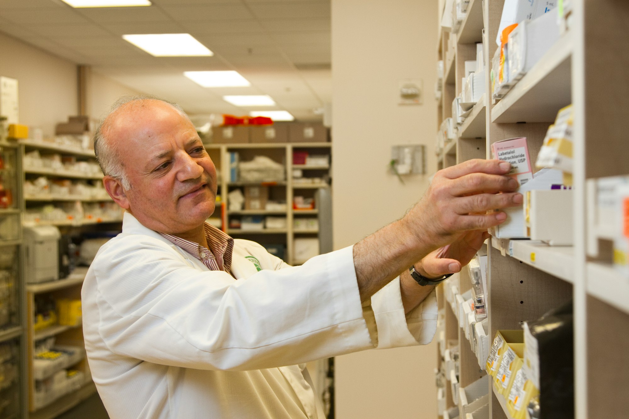 A male pharmacist is selecting a drug from a display case in a pharmacy.