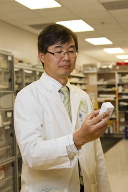 A person wearing a white lab coat and glasses is holding a small box, standing in a room that appears to be a laboratory or storage area for scientific or medical supplies. Shelves with various items are visible in the background.