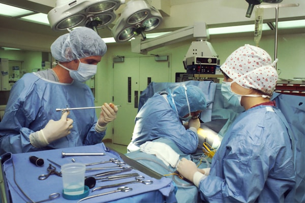 Medical professionals wearing surgical gowns and masks are engaged in a surgical procedure in an operating room. Various surgical instruments are neatly arranged on a blue surgical drape. Overhead surgical lights illuminate the scene, and medical equipment is visible in the background.