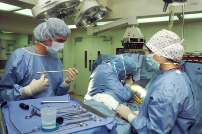 Medical professionals wearing surgical gowns and masks are engaged in a surgical procedure in an operating room. Various surgical instruments are neatly arranged on a blue surgical drape. Overhead surgical lights illuminate the scene, and medical equipment is visible in the background.