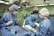 Medical professionals wearing surgical gowns and masks are engaged in a surgical procedure in an operating room. Various surgical instruments are neatly arranged on a blue surgical drape. Overhead surgical lights illuminate the scene, and medical equipment is visible in the background.