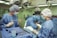 Medical professionals wearing surgical gowns and masks are engaged in a surgical procedure in an operating room. Various surgical instruments are neatly arranged on a blue surgical drape. Overhead surgical lights illuminate the scene, and medical equipment is visible in the background.