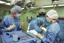 Medical professionals wearing surgical gowns and masks are engaged in a surgical procedure in an operating room. Various surgical instruments are neatly arranged on a blue surgical drape. Overhead surgical lights illuminate the scene, and medical equipment is visible in the background.