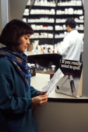 A woman wearing a blue sweater and scarf is looking at a piece of paper with a drawing on it. In the background, a pharmacist in a white coat is working behind the counter, which is filled with numerous medicine bottles. A sign on the counter reads 'If you really want to quit.'