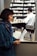 A woman wearing a blue sweater and scarf is looking at a piece of paper with a drawing on it. In the background, a pharmacist in a white coat is working behind the counter, which is filled with numerous medicine bottles. A sign on the counter reads 'If you really want to quit.'