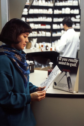 A woman wearing a blue sweater and scarf is looking at a piece of paper with a drawing on it. In the background, a pharmacist in a white coat is working behind the counter, which is filled with numerous medicine bottles. A sign on the counter reads 'If you really want to quit.'