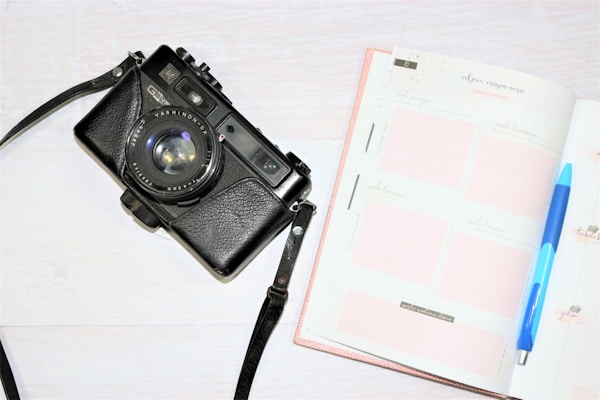 A vintage-style black camera with a large lens is positioned on the left side. To the right, there is an open planner with blank pages displayed, along with a blue pen resting on top.