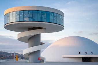 A modern architectural structure featuring a circular building with large windows, elevated above the ground on a cylindrical pillar. A spiral ramp encircles the pillar, providing access to the structure. Adjacent to this is a large, dome-shaped building with a sleek white surface and small circular windows.