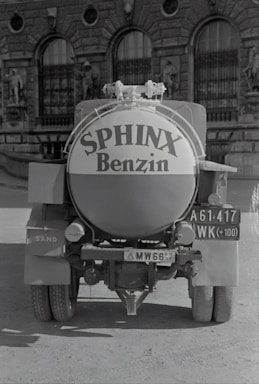A vintage fuel truck with a cylindrical tank displays the words 'Sphinx Benzin' prominently on its back. The vehicle has classic, rugged tires and is parked on a dirt road. Behind it stands an ornate building with arched windows and statues, suggesting an older architectural style. The truck carries a license plate with various numbers and letters.