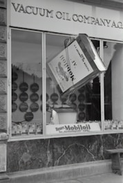 A black and white image of a storefront with the sign 'VACUUM OIL COMPANY A.G.' prominently displayed at the top. In the center of a large window is a large model of a can labeled 'Mobiloil', which appears to be part of an elaborate display. The display within the window includes various circular elements and additional cans arranged neatly along the bottom edge. The overall scene has a vintage and historical feel.