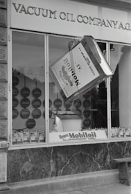 A black and white image of a storefront with the sign 'VACUUM OIL COMPANY A.G.' prominently displayed at the top. In the center of a large window is a large model of a can labeled 'Mobiloil', which appears to be part of an elaborate display. The display within the window includes various circular elements and additional cans arranged neatly along the bottom edge. The overall scene has a vintage and historical feel.