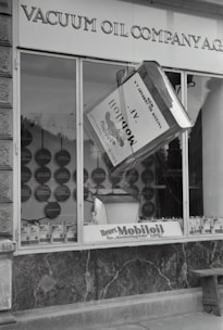 A black and white image of a storefront with the sign 'VACUUM OIL COMPANY A.G.' prominently displayed at the top. In the center of a large window is a large model of a can labeled 'Mobiloil', which appears to be part of an elaborate display. The display within the window includes various circular elements and additional cans arranged neatly along the bottom edge. The overall scene has a vintage and historical feel.