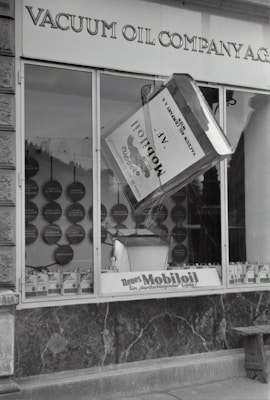 A black and white image of a storefront with the sign 'VACUUM OIL COMPANY A.G.' prominently displayed at the top. In the center of a large window is a large model of a can labeled 'Mobiloil', which appears to be part of an elaborate display. The display within the window includes various circular elements and additional cans arranged neatly along the bottom edge. The overall scene has a vintage and historical feel.