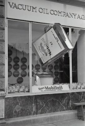 A black and white image of a storefront with the sign 'VACUUM OIL COMPANY A.G.' prominently displayed at the top. In the center of a large window is a large model of a can labeled 'Mobiloil', which appears to be part of an elaborate display. The display within the window includes various circular elements and additional cans arranged neatly along the bottom edge. The overall scene has a vintage and historical feel.
