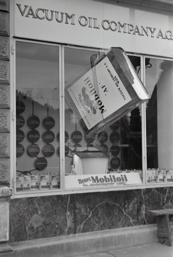 A black and white image of a storefront with the sign 'VACUUM OIL COMPANY A.G.' prominently displayed at the top. In the center of a large window is a large model of a can labeled 'Mobiloil', which appears to be part of an elaborate display. The display within the window includes various circular elements and additional cans arranged neatly along the bottom edge. The overall scene has a vintage and historical feel.