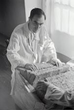 A person in a lab coat examines a large rectangular block composed of layered material. The setting appears to be a dimly-lit room with light coming from a window. The person's hands rest on the surface of the block, and another person's hands are visible on the opposite side.