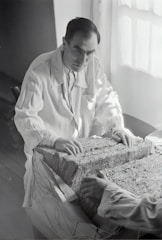 A person in a lab coat examines a large rectangular block composed of layered material. The setting appears to be a dimly-lit room with light coming from a window. The person's hands rest on the surface of the block, and another person's hands are visible on the opposite side.