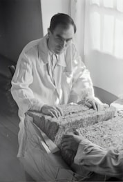 A person in a lab coat examines a large rectangular block composed of layered material. The setting appears to be a dimly-lit room with light coming from a window. The person's hands rest on the surface of the block, and another person's hands are visible on the opposite side.