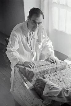 A person in a lab coat examines a large rectangular block composed of layered material. The setting appears to be a dimly-lit room with light coming from a window. The person's hands rest on the surface of the block, and another person's hands are visible on the opposite side.