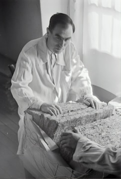 A person in a lab coat examines a large rectangular block composed of layered material. The setting appears to be a dimly-lit room with light coming from a window. The person's hands rest on the surface of the block, and another person's hands are visible on the opposite side.