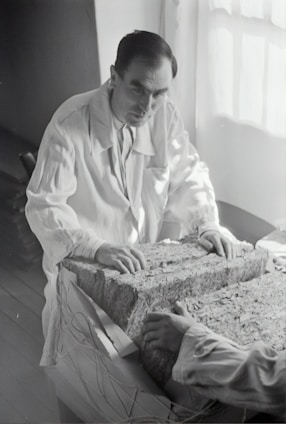 A person in a lab coat examines a large rectangular block composed of layered material. The setting appears to be a dimly-lit room with light coming from a window. The person's hands rest on the surface of the block, and another person's hands are visible on the opposite side.