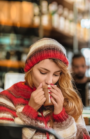 A woman with long hair is sipping a hot beverage from a mug. She is wearing a striped knit beanie and a matching sweater in shades of red, cream, and brown. The background is a blurred interior, possibly a café, with shelves and warm lighting.