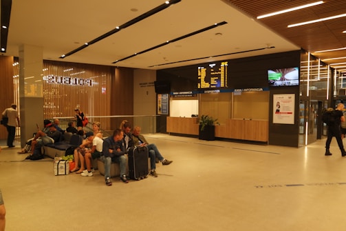 A modern indoor waiting area with several people seated on benches, some with luggage. A large display board shows travel information above a kiosk with ticket counters. The space is well-lit with wooden panels and minimalistic design.