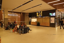 A modern indoor waiting area with several people seated on benches, some with luggage. A large display board shows travel information above a kiosk with ticket counters. The space is well-lit with wooden panels and minimalistic design.