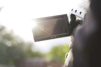A person is holding a video camera with a focus on the camera's screen which is partially illuminated by bright sunlight. The background is out of focus, showing some greenery and creating a soft, serene atmosphere.