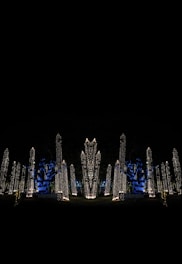 Tall structures are adorned with a multitude of tiny, warm white lights against a dark background. Blue uplighting highlights the trees, creating a vibrant contrast with the gold-colored lights. The scene suggests artificial illumination in a nighttime outdoor setting.