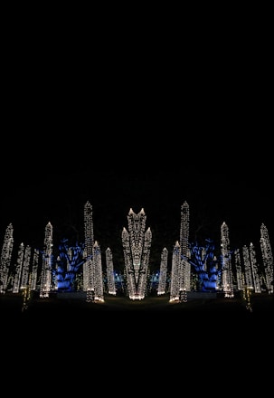 Tall structures are adorned with a multitude of tiny, warm white lights against a dark background. Blue uplighting highlights the trees, creating a vibrant contrast with the gold-colored lights. The scene suggests artificial illumination in a nighttime outdoor setting.