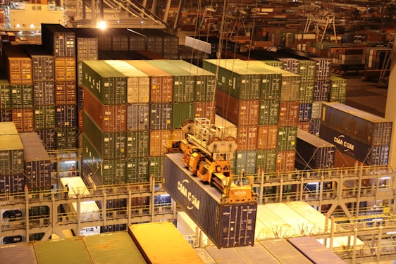 An overhead view of a bustling port with numerous stacked shipping containers in various colors. A large crane is actively loading or unloading a container labeled 'CMA CGM'. The area is illuminated with artificial lighting, casting warm yellow tones across the scene, typical of night operations.