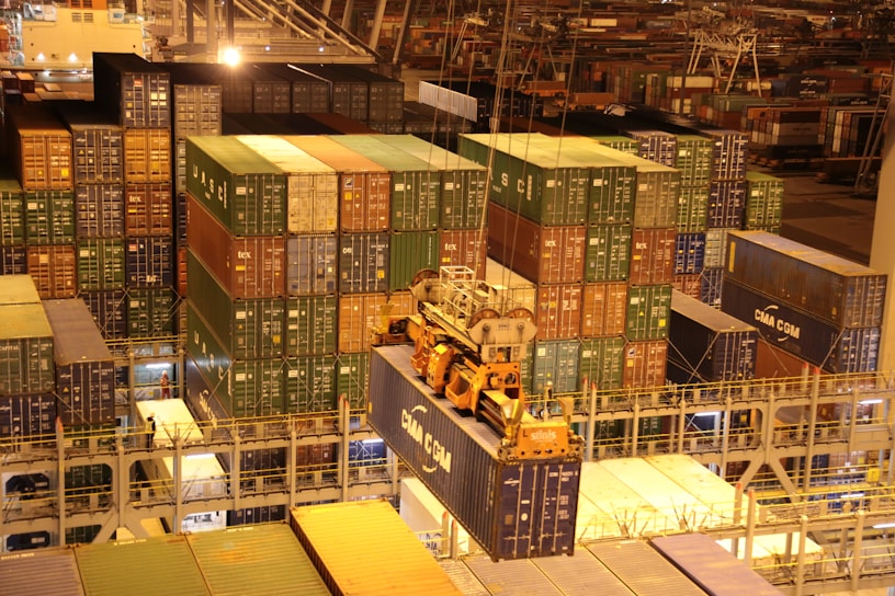 An overhead view of a bustling port with numerous stacked shipping containers in various colors. A large crane is actively loading or unloading a container labeled 'CMA CGM'. The area is illuminated with artificial lighting, casting warm yellow tones across the scene, typical of night operations.