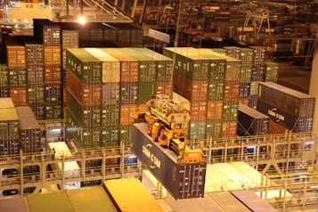 An overhead view of a bustling port with numerous stacked shipping containers in various colors. A large crane is actively loading or unloading a container labeled 'CMA CGM'. The area is illuminated with artificial lighting, casting warm yellow tones across the scene, typical of night operations.