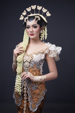 A woman is wearing traditional Indonesian attire featuring intricate embroidery and embellishments in white, gold, and silver tones. Her outfit is accented with a large floral and lace applique on the shoulder. She has an ornate headdress adorned with floral decorations and a long braid adorned with jasmine flowers flowing down her front. Her makeup is elegant, highlighting her eyes and lips.