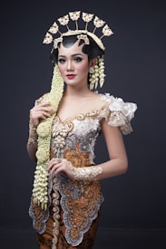 A woman is wearing traditional Indonesian attire featuring intricate embroidery and embellishments in white, gold, and silver tones. Her outfit is accented with a large floral and lace applique on the shoulder. She has an ornate headdress adorned with floral decorations and a long braid adorned with jasmine flowers flowing down her front. Her makeup is elegant, highlighting her eyes and lips.