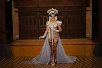 A person wearing a traditional outfit with elaborate decorative elements, including a headdress and a gown with ornate patterns. The background includes wooden carvings and a patterned floor.