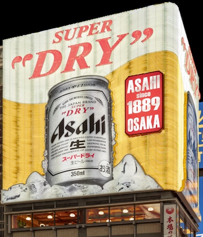 A large illuminated billboard displays a can of Asahi Super Dry beer, prominently featuring its brand name and various text in English and Japanese. The can is shown against a backdrop of ice cubes, emphasizing coldness and refreshment, with additional text indicating the beer's history since 1889 in Osaka.