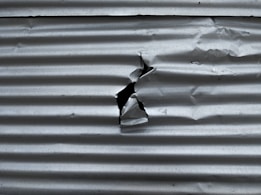 A sheet of corrugated metal with a noticeable tear in the center. The surface exhibits a series of parallel horizontal ridges, common in metal sheeting used for construction or roofing.