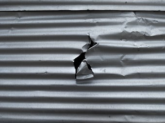 A sheet of corrugated metal with a noticeable tear in the center. The surface exhibits a series of parallel horizontal ridges, common in metal sheeting used for construction or roofing.