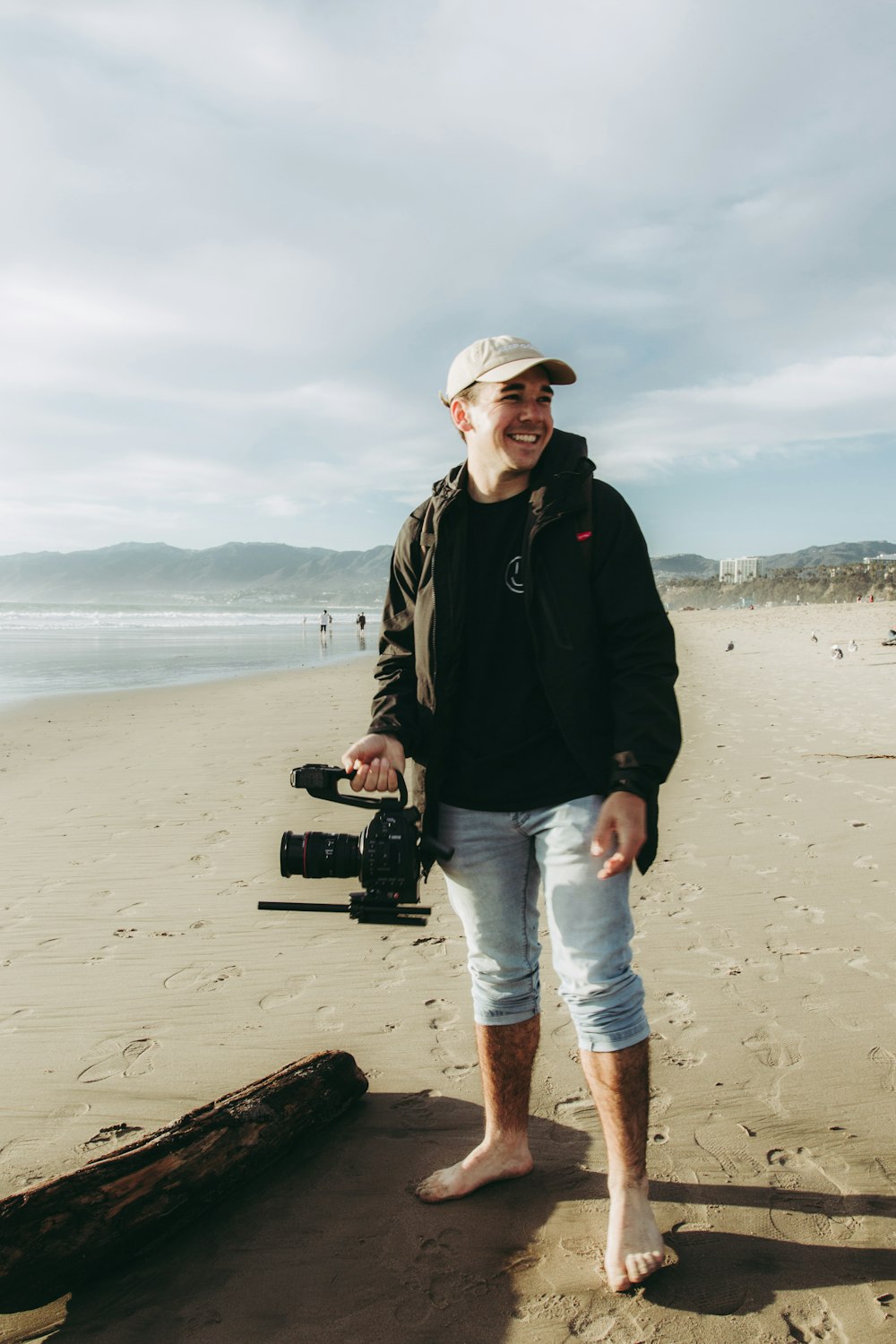 Mann in schwarzer Jacke und blauer Jeans mit schwarzer DSLR-Kamera am Strand während