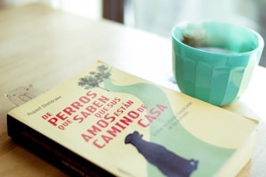 A book with a beige cover and Spanish text is lying on a wooden table. Next to it, there is a teal cup, filled with a hot beverage. The table has a warm, natural tone, and the background is softly blurred.