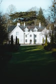 A large, elegant white house with multiple peaked roofs and chimneys, surrounded by tall trees and lush greenery. The house features several windows with white frames and a central front door. Manicured lawns and hedges lead up to the entrance, creating a grandiose and tranquil atmosphere.