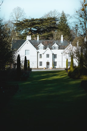 A large, elegant white house with multiple peaked roofs and chimneys, surrounded by tall trees and lush greenery. The house features several windows with white frames and a central front door. Manicured lawns and hedges lead up to the entrance, creating a grandiose and tranquil atmosphere.