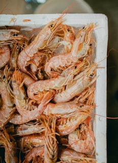 A collection of fresh shrimps is packed closely together inside a light-colored container. The shrimps are pinkish-orange with visible eyes and antennae, creating a textured and detailed scene.