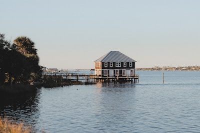 A lake house with a metal roof sits on stilts above the water. Surrounding the house are calm waters reflecting the early morning or late afternoon light. On the left, trees and vegetation line the shoreline, while more houses and structures are visible in the distance across the water.