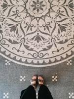Ornate black and white mosaic floor design featuring intricate floral and geometric patterns at the top. A pair of feet in sandals with dark pants are visible standing on this detailed flooring.
