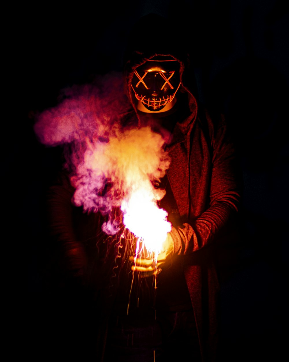 Person in schwarzer Maske, die lila und rosa Rauch hält